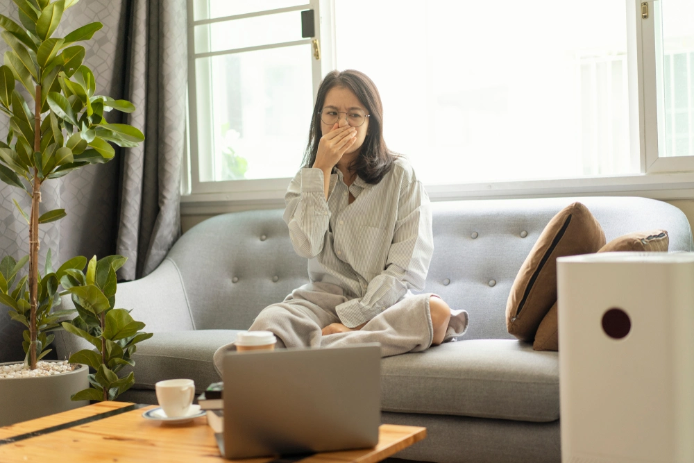 A woman sitting on a couch, covering her nose with a grimace.