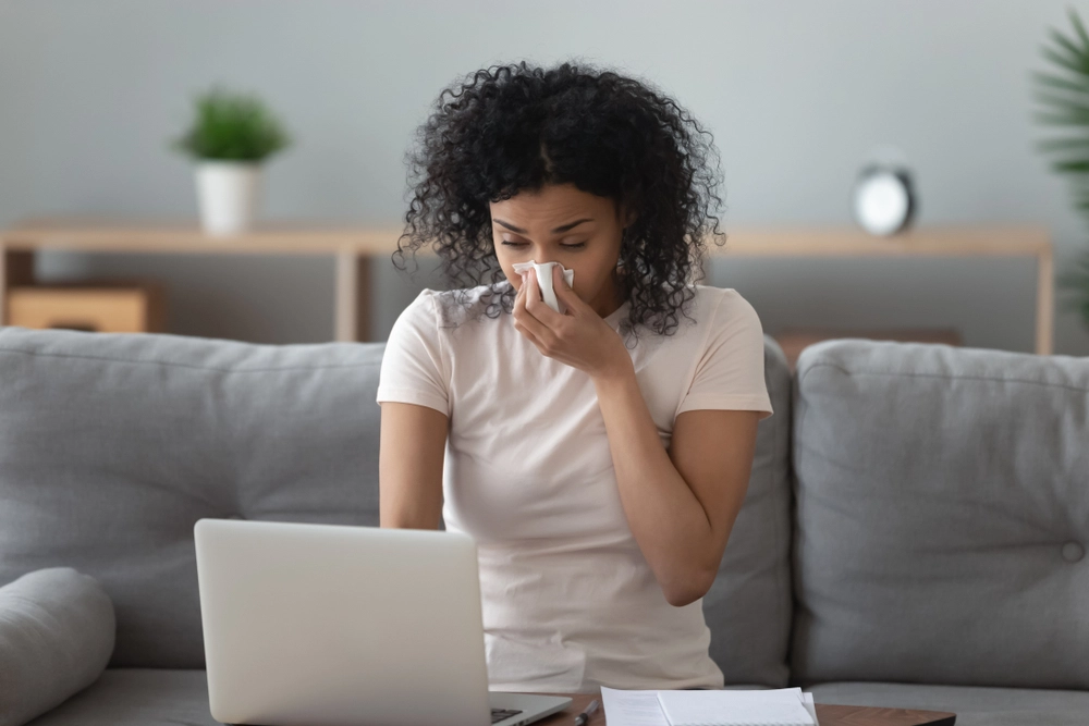 Woman sneezes into tissue while looking on laptop.
