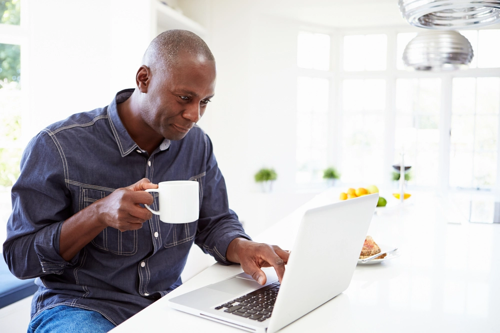 Smiling man with mug in his hand sits at table while looking laptop.