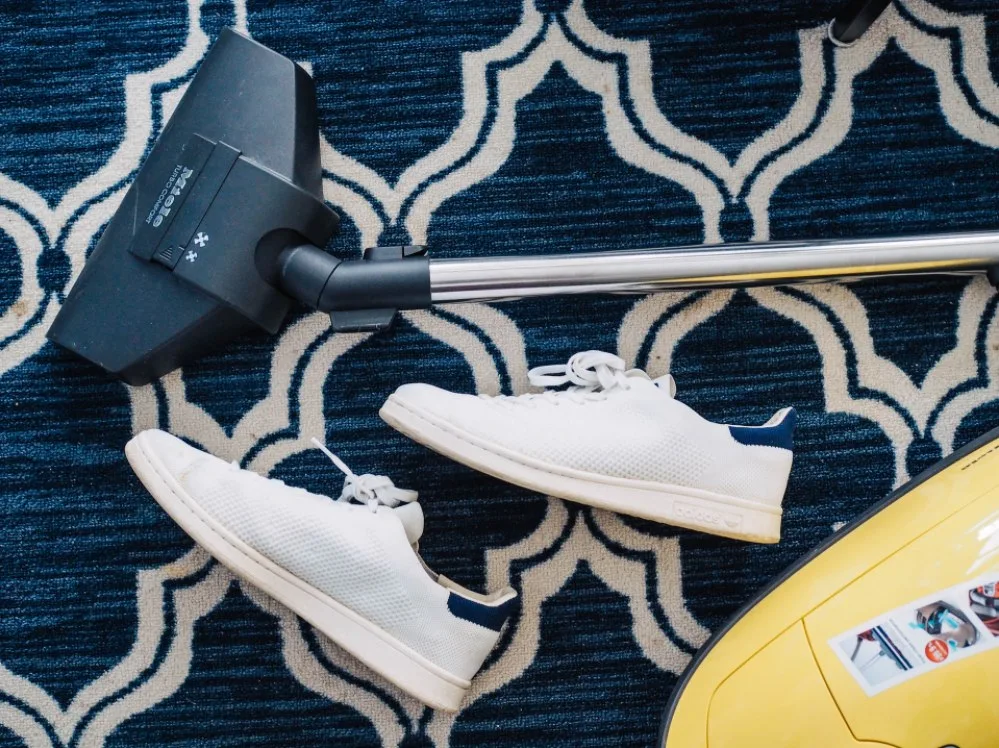 A grey vacuum and white shoes sit on a blue and white carpet.
