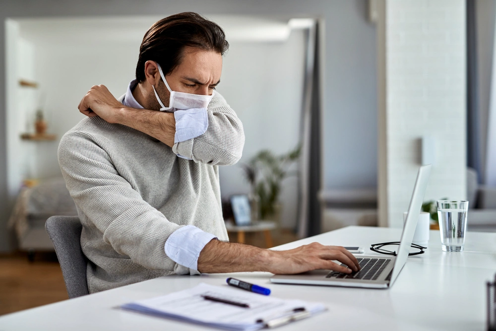 Man working at computer covers his mouth while coughing.
