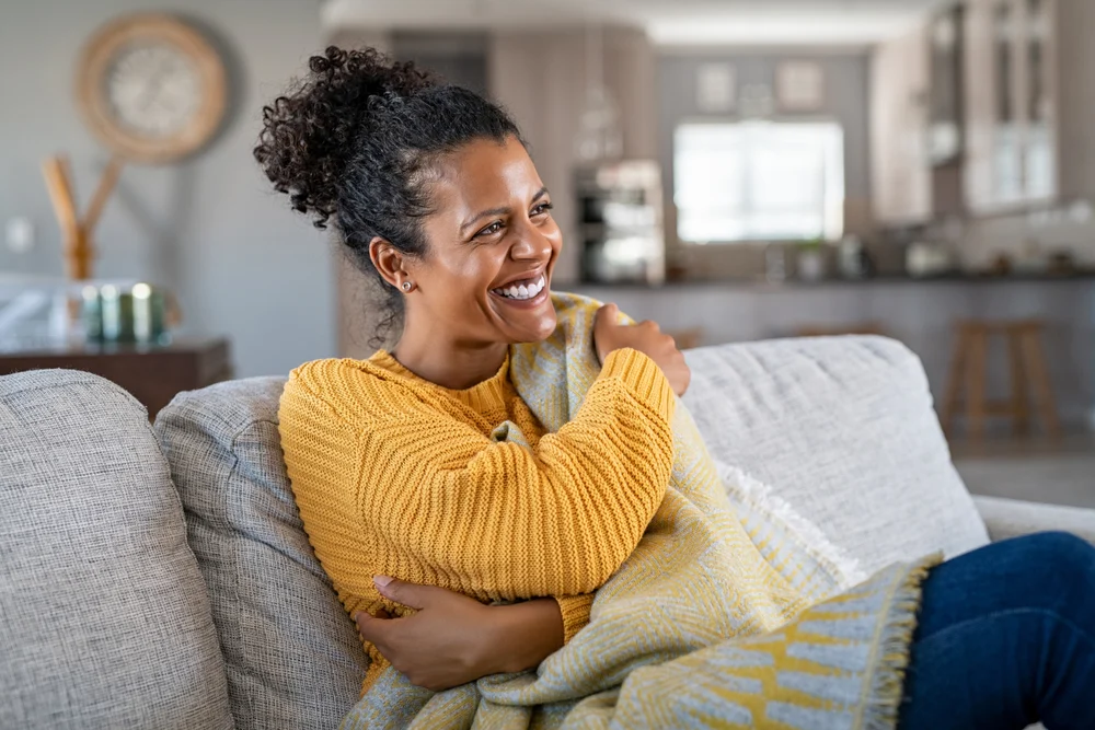 Smiling women sits on her couch wrapped in a blanket.