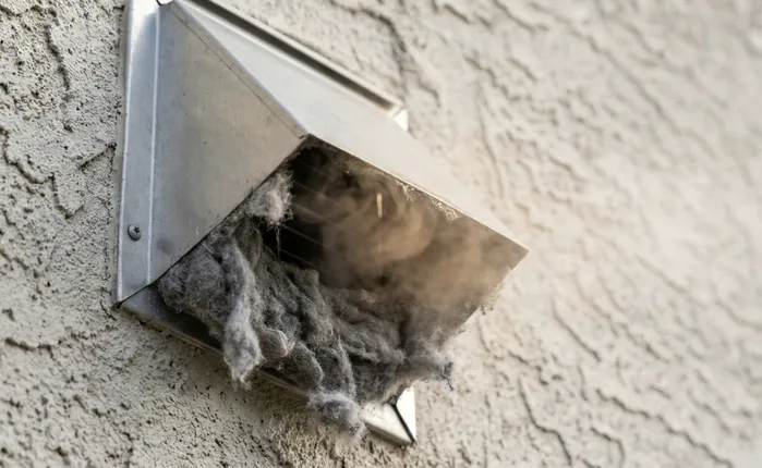 An exterior dryer vent hood is shown completely blocked by a large mass of gray lint, with steam escaping from the restricted opening, illustrating common clogged dryer vent symptoms such as excessive lint accumulation and reduced airflow.